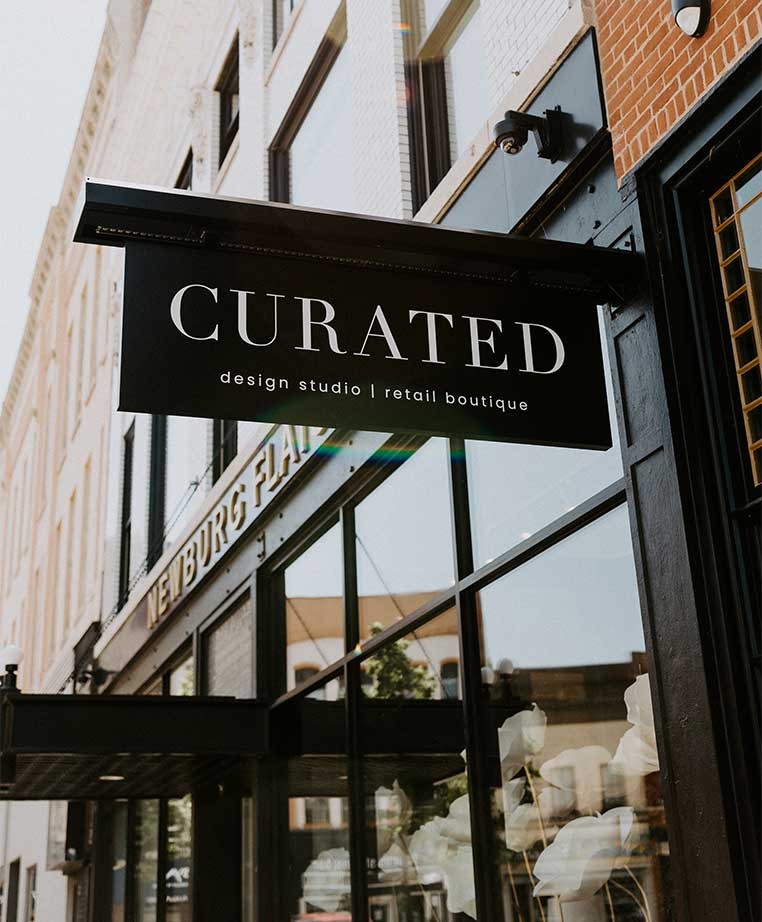 A black sign hanging from a brick building with the caption "Curated, design studio and retail boutique"